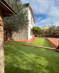 Jardin moderne avec pelouse synthétique, terrasse en bois rouge, mur en pierre d'une maison et olivier sous un ciel bleu nuageux.