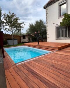 Homme joyeux fait un geste de pouce levé devant une piscine rectangulaire moderne avec terrasse en bois rouge, près d'une maison blanche.