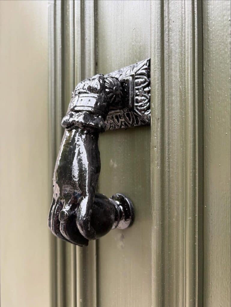 Gros plan sur une poignée de porte en laiton noirci, en forme de main fermée, sur une porte en bois vert olive à rainures verticales.