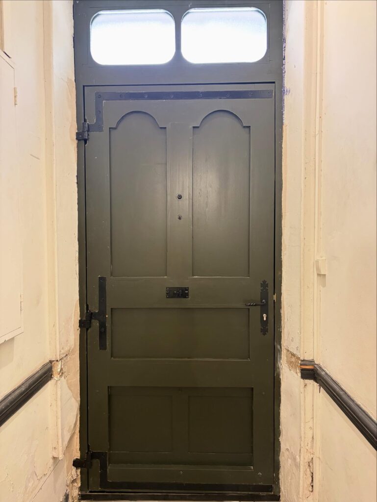 Porte d'entrée ancienne, peinte en vert foncé, avec fenêtres en verre dépoli en partie haute et ferronnerie noire robuste.