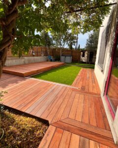 Terrasse en bois rouge moderne avec un panneau coulissant couvrant une piscine ou un espace de vie, juxtaposée à une pelouse synthétique verte.