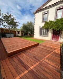 Grande terrasse en bois exotique rougeâtre avec un panneau rétractable dissimulant potentiellement une piscine ou un jacuzzi, devant une maison traditionnelle.
