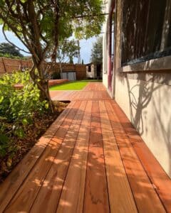 Terrasse en bois rouge neuf bordant un mur clair, avec ombre d'arbre filtrant la lumière du soleil, menant vers une pelouse.