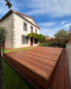 Grande terrasse surélevée en bois rouge-brun neuf devant une maison ancienne aux murs blancs et pierre, sous un ciel bleu nuageux.