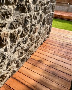 Gros plan sur une terrasse en bois rouge-brun posée contre un mur ancien en pierres brutes et mortier clair, sous un soleil éclatant.